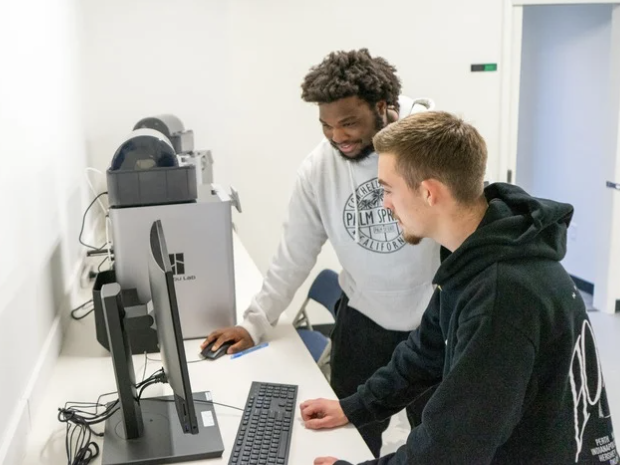 Two men work together at a computer station in a lab, both looking at the screen and interacting with the keyboard and mouse.